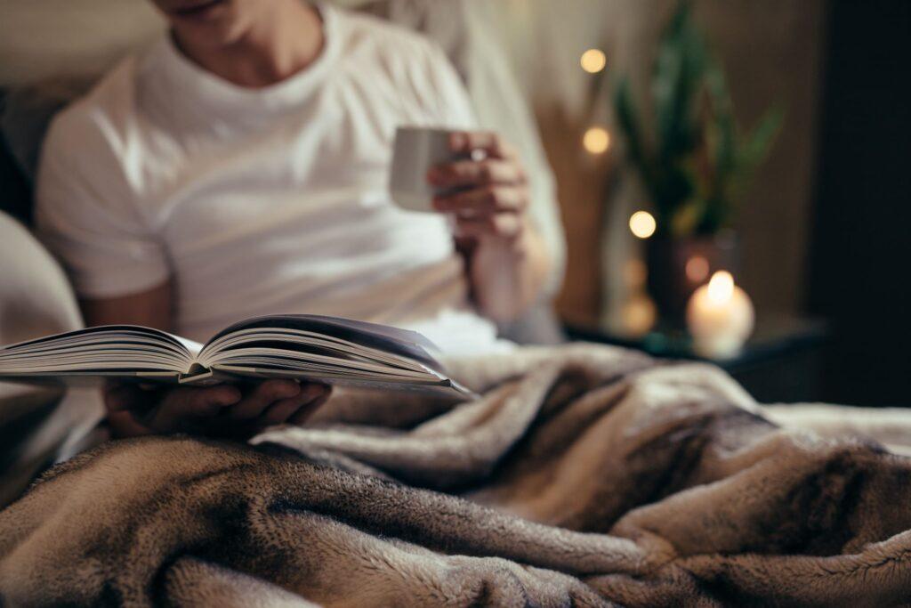 Eine Person liest ein Buch und hält eine Tasse Kaffee, eingewickelt in eine weiche, warme Alpaka Decke. Im Hintergrund sorgen Kerzen und Pflanzen für eine behagliche Atmosphäre. 