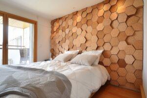 Ein stilvoll gestaltetes Schlafzimmer mit einer Wand aus hexagonalen Korkplatten. Die Korkwand verleiht dem Raum eine warme und natürliche Atmosphäre. Das Bett ist mit weißen Kissen und einer grauen Decke dekoriert. Große Fenster lassen viel natürliches Licht herein, das die Struktur und Farbe der Korkwand betont.