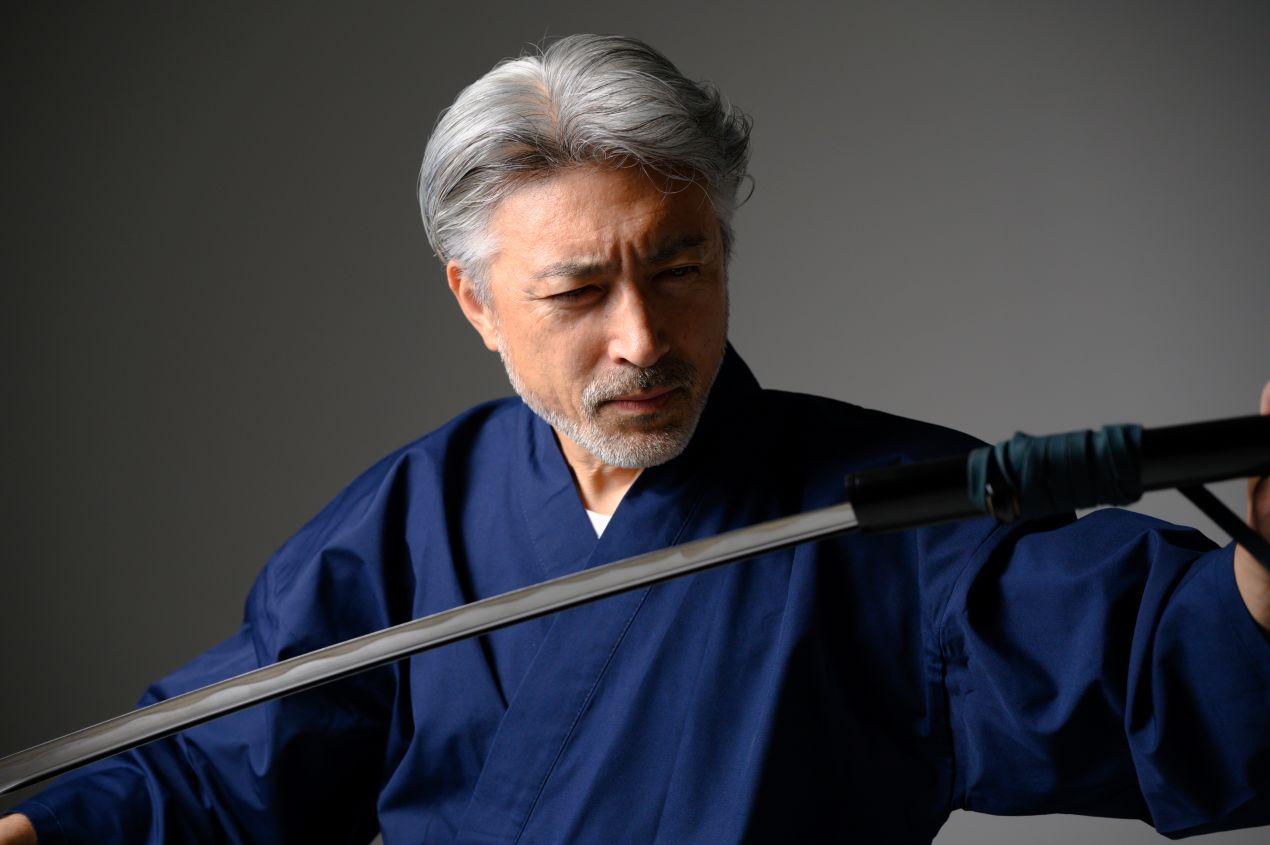 Älterer Mann im traditionellen blauen Gewand betrachtet ein Katana sorgfältig, Symbol für japanische Handwerkskunst und Samurai-Tradition.