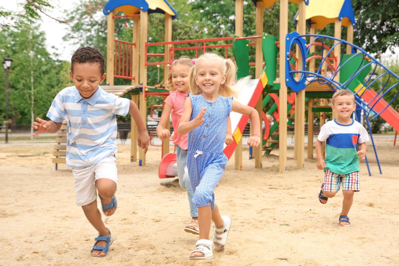 Vier fröhliche Kinder rennen über einen Spielplatz mit bunten Spielplatzgeräten. Perfekte Umgebung für Bewegung, Abenteuer und soziale Interaktion.