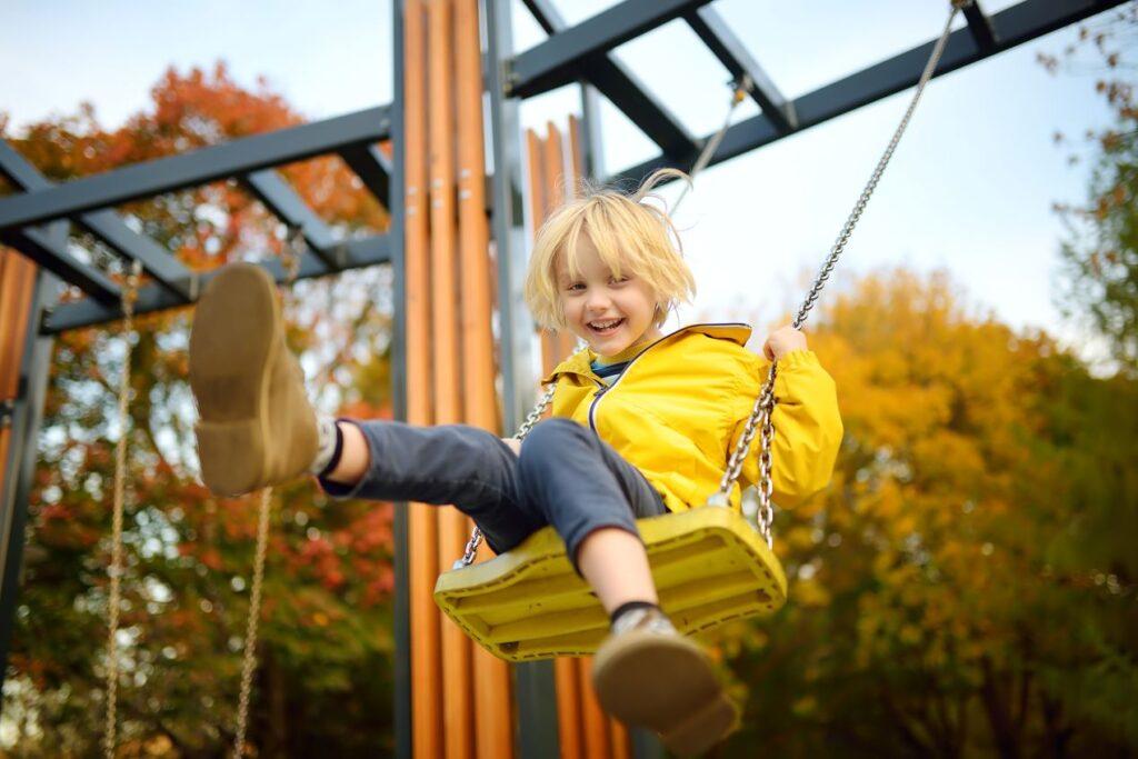 Ein lachendes Kind mit gelber Jacke schaukelt auf einem modernen Spielplatz. Hochwertige Spielplatzgeräte für sicheres Spielen und Bewegung.