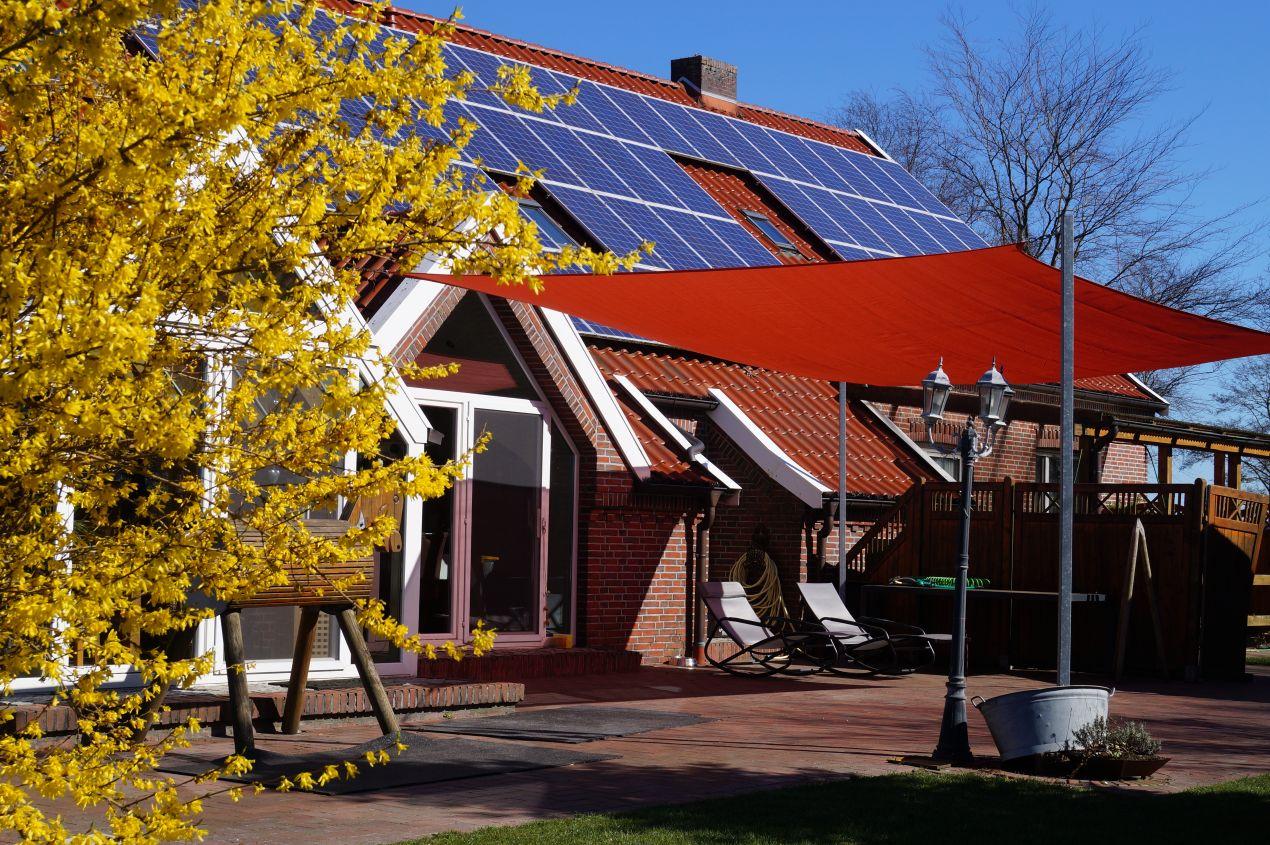 Sonnensegelmast mit rotem Segel auf Terrasse eines Hauses mit Solarpanelen und gelbem Fruehlingsbaum